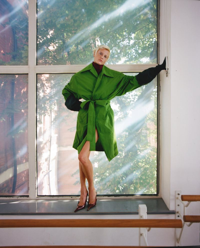 A fashion shot of a woman in a green coat and black gloves posing in front of a window with natural light, showcasing editorial style.
