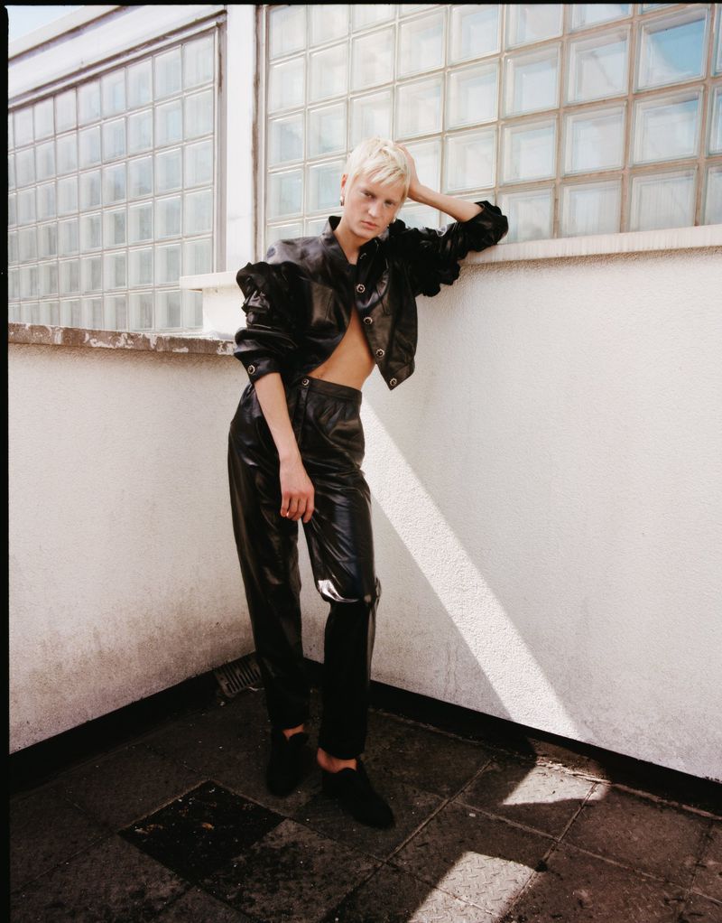 An adult woman poses leaning against a wall wearing a black leather jacket and pants with a glass-block window in the background.