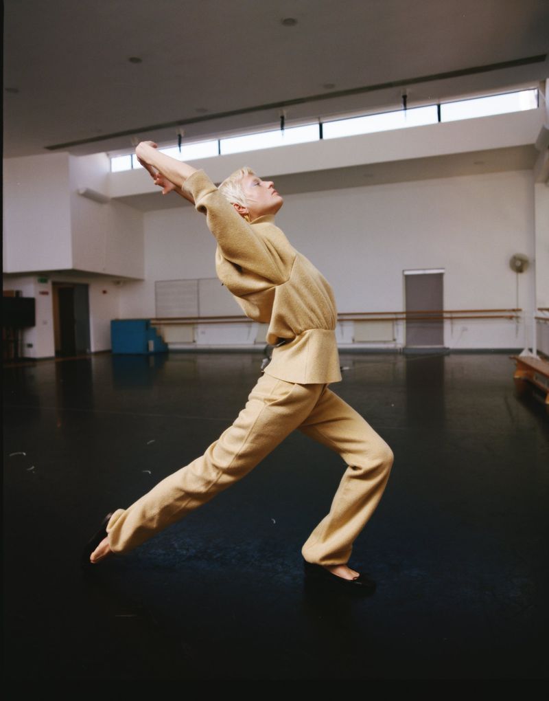Adult female in a tan two-piece outfit in a dance pose in a studio with black floor and white walls and natural light from upper windows.
