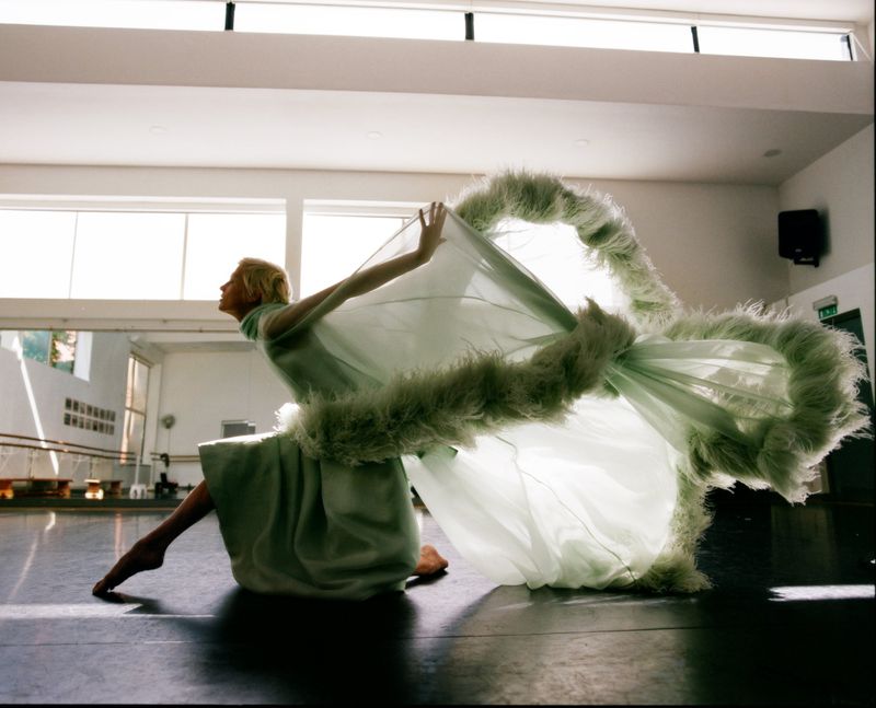 Elegant woman in a light green dress poses dynamically indoors, backlit, casting shadows on the dark floor of a spacious, bright dance studio.