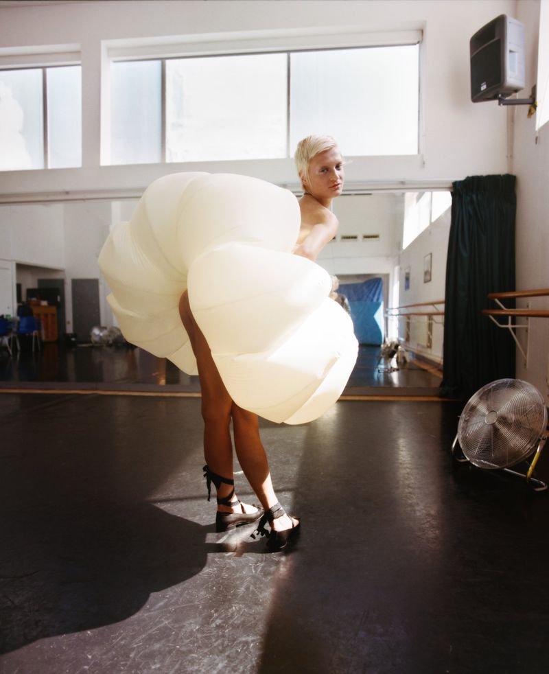 Woman with short hair and high heels in a white voluminous dress in a dance studio with ballet barres and large windows.
