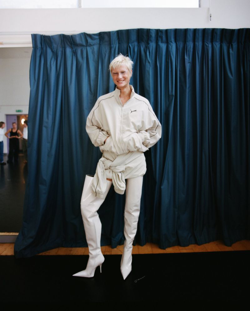 Woman in a light-colored tracksuit and white thigh-high boots poses against a blue curtain in a photo studio setting with a confident smile.