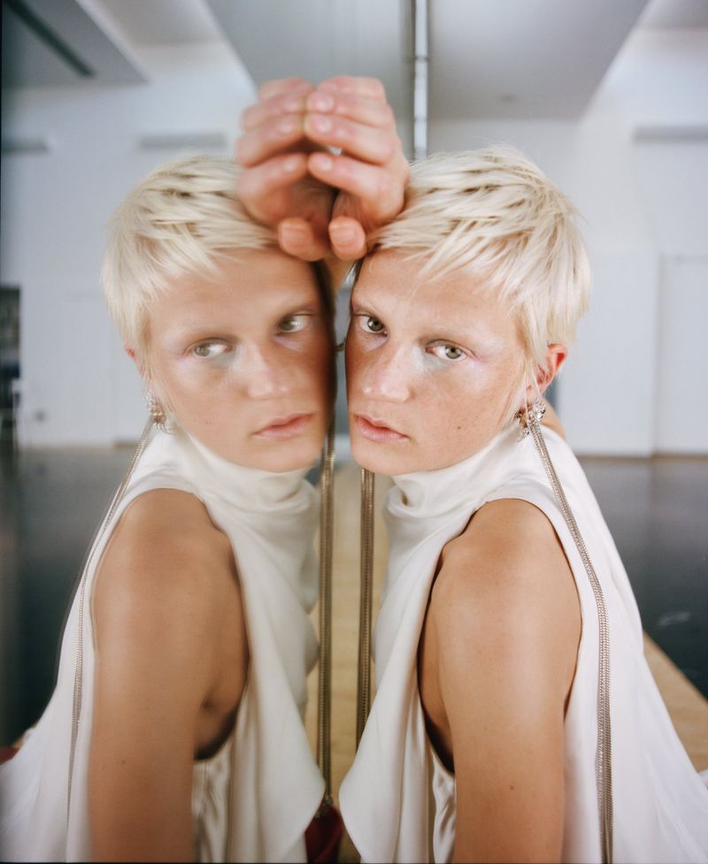 Close-up of a person with platinum blonde hair looking into a mirror with hands on the glass, wearing a sleeveless white top.