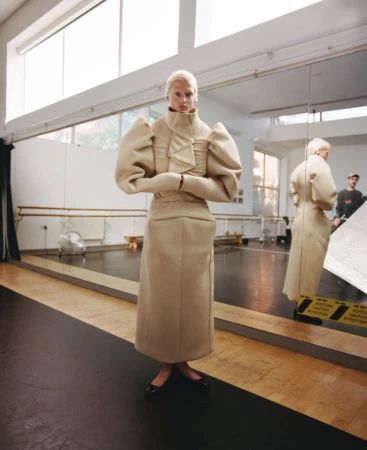 Blonde woman poses in an oversized beige outfit in a dance studio, reflecting in a mirror, capturing a fashion editorial shot.