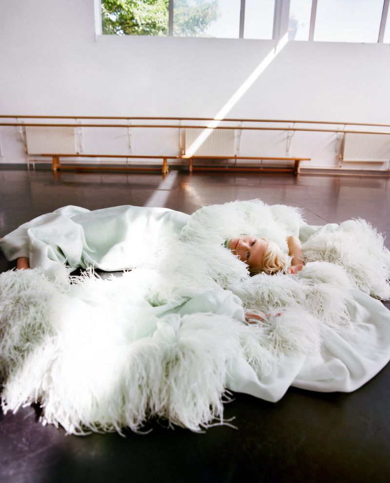 Woman in flowing feathered white gown reclines on a dark floor in a bright room with barre. Natural light stream in through windows.