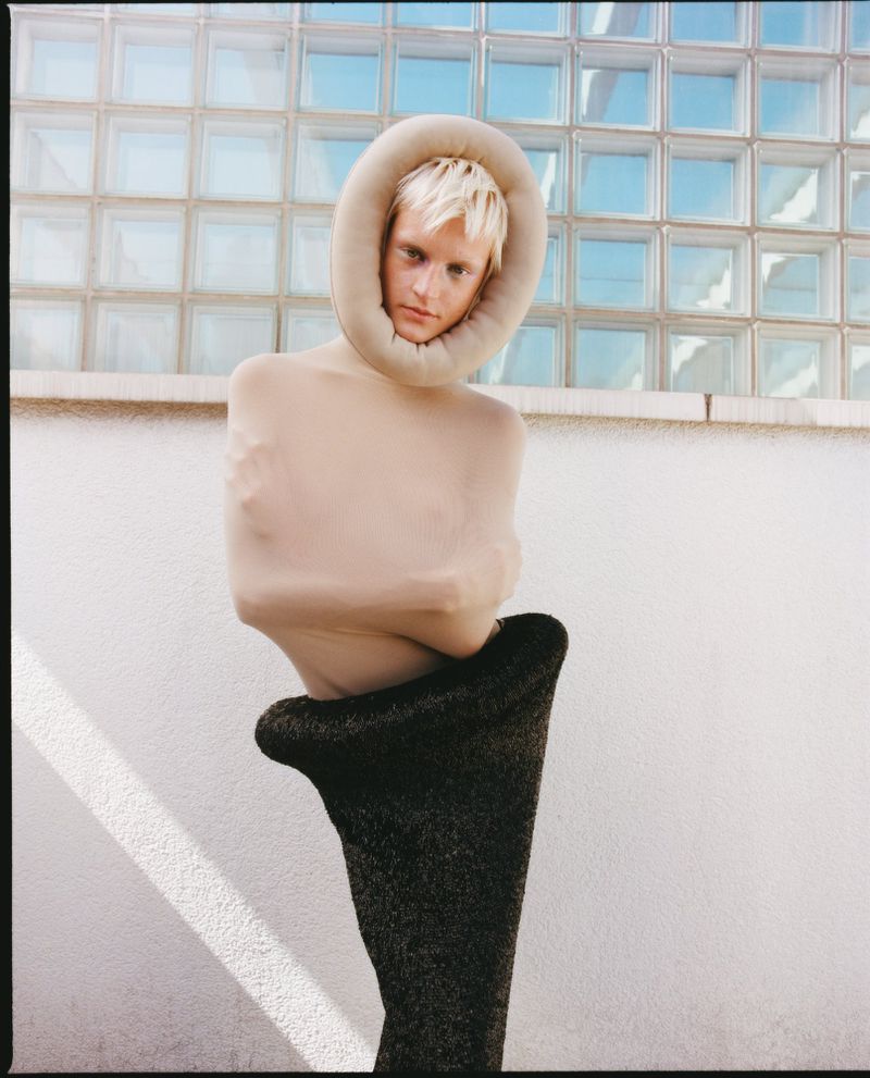 Unique fashion image featuring an adult with blonde hair wearing an avant-garde tan neckpiece and dark grey form fitting garment outdoors.