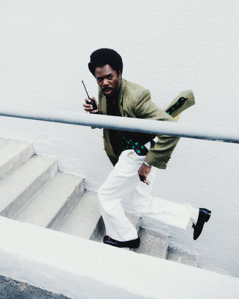 An African-American man is running down a flight of stairs while holding a walkie-talkie, dressed in retro clothing, on a white outdoor staircase.