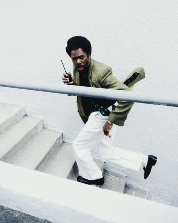 An African-American man is running down a flight of stairs while holding a walkie-talkie, dressed in retro clothing, on a white outdoor staircase.