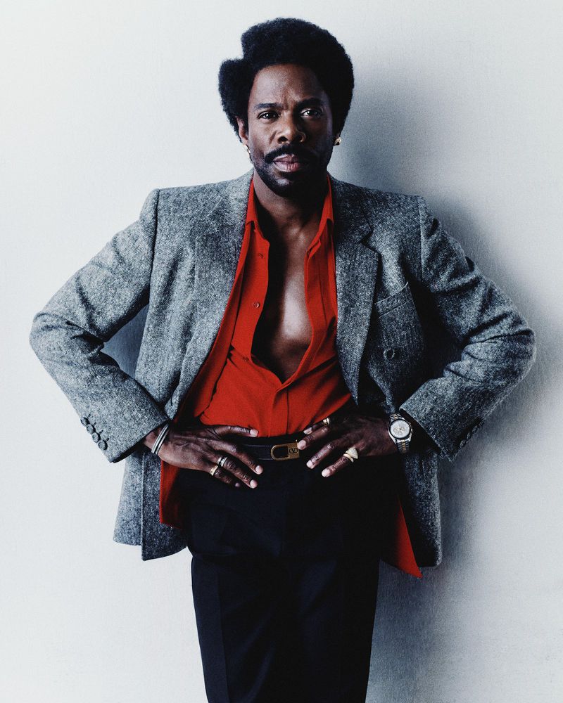 Portrait of an adult Black male wearing a gray blazer and red shirt with his hands on his hips against a white background.