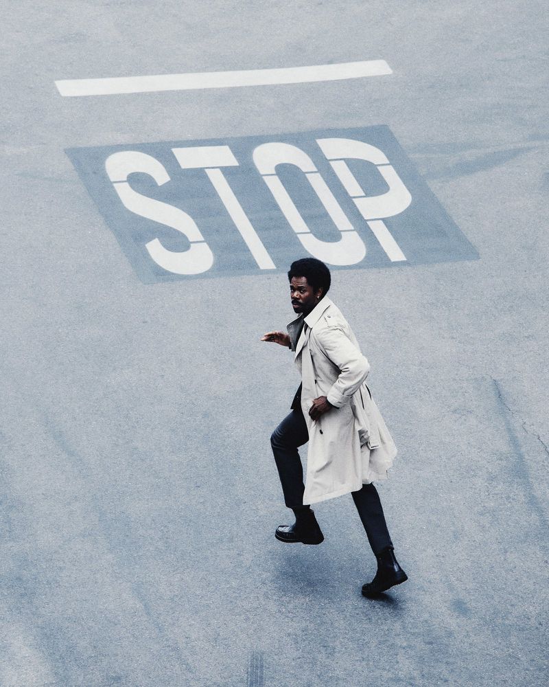 Aerial view of an adult male running across a concrete surface marked with a large stenciled "STOP" sign, wearing a trench coat.