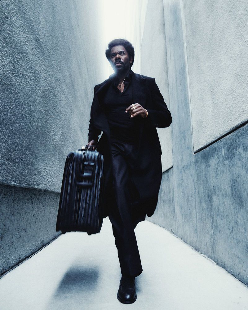 A sharply dressed man runs confidently through a modern urban space with a structured black carry-on suitcase, bathed in diffused light.