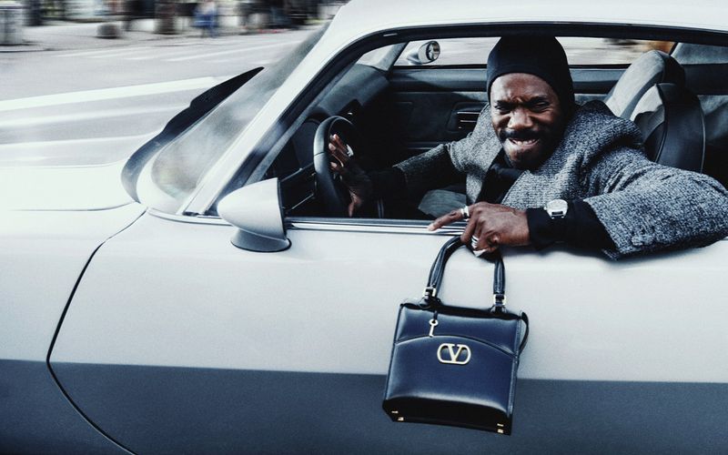 Adult male driving a white car, wearing a black hat and grey coat, carrying a black leather Valentino handbag in an urban setting.