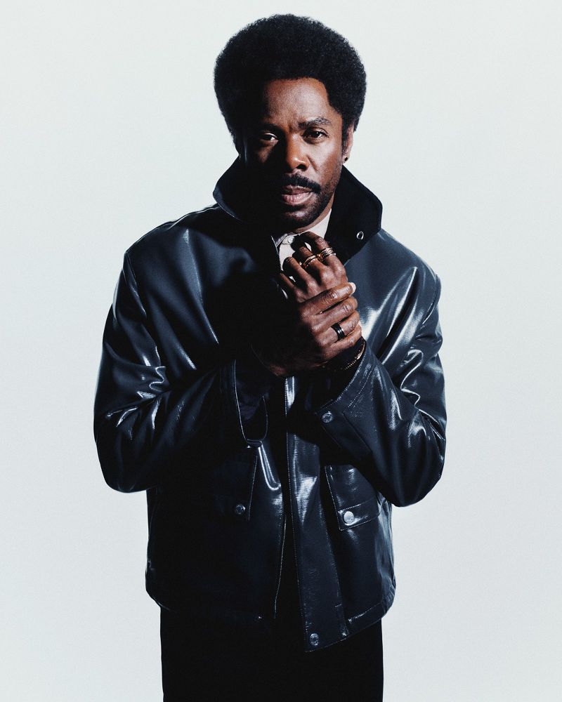 Actor Colman Domingo wearing a glossy black jacket and gold rings, posing against a white seamless backdrop.
