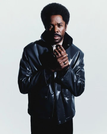 Actor Colman Domingo wearing a glossy black jacket and gold rings, posing against a white seamless backdrop.