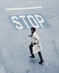 Colman Domingo running across an asphalt street marked with a painted stop sign in a beige trench coat and dark trousers.