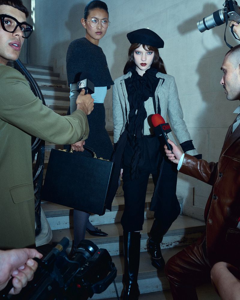 Fashion scene with a woman in a beret and gray jacket being interviewed by reporters holding microphones on a staircase indoors.