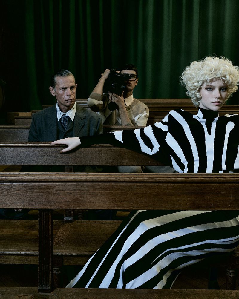 Editorial fashion shot of a model in a black and white striped dress posing near wooden pews, with two men in the background.