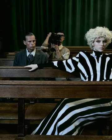 Editorial fashion shot of a model in a black and white striped dress posing near wooden pews, with two men in the background.