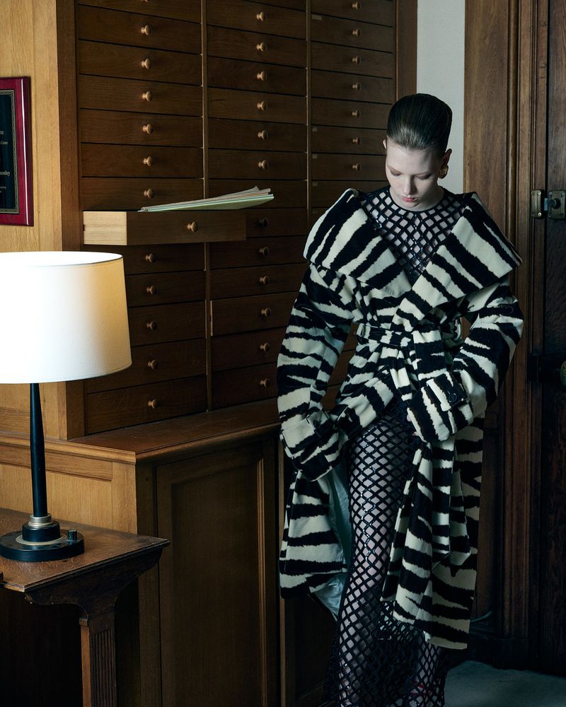 A woman poses in a black and white coat with fishnet leggings in an indoor environment with neutral-toned wooden walls and furniture.