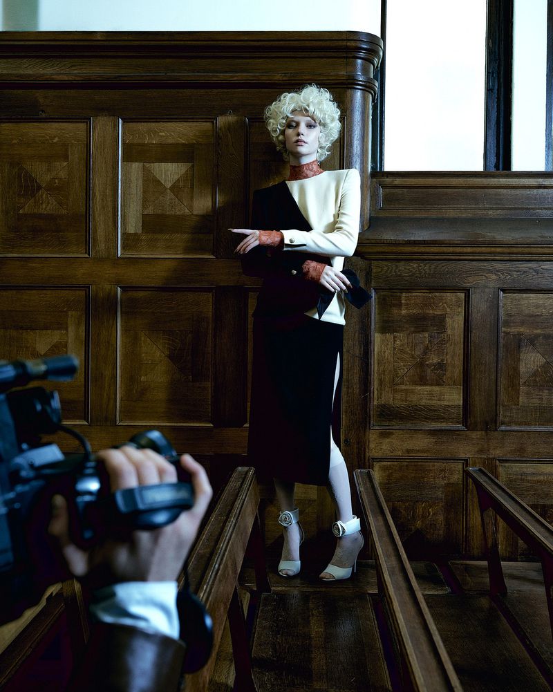 A woman in a two-tone dress poses, photographed by a hand-held camera in an indoor setting with wood paneling and flooring.