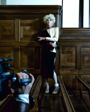 A woman in a two-tone dress poses, photographed by a hand-held camera in an indoor setting with wood paneling and flooring.