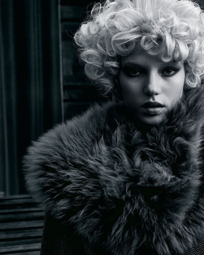 Fashionable adult woman with curly blonde hair wearing a fur collar. Indoor studio setting with dark background. Black and white portrait.