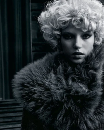 Fashionable adult woman with curly blonde hair wearing a fur collar. Indoor studio setting with dark background. Black and white portrait.