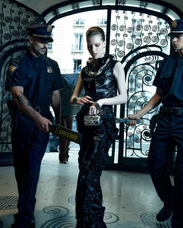 Fashion model in a black dress is scanned by security guards holding metal detectors; ornate wrought iron gate and city background.