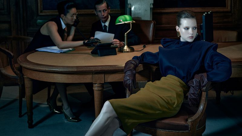 Fashion photography featuring a woman in a blue top and yellow skirt sitting in a chair in an office, with two people at a desk in the background.