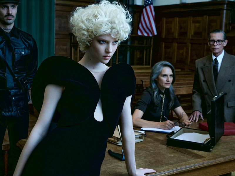 Fashion editorial image featuring a blonde woman in a black dress, seated older woman writing, and an American flag backdrop.