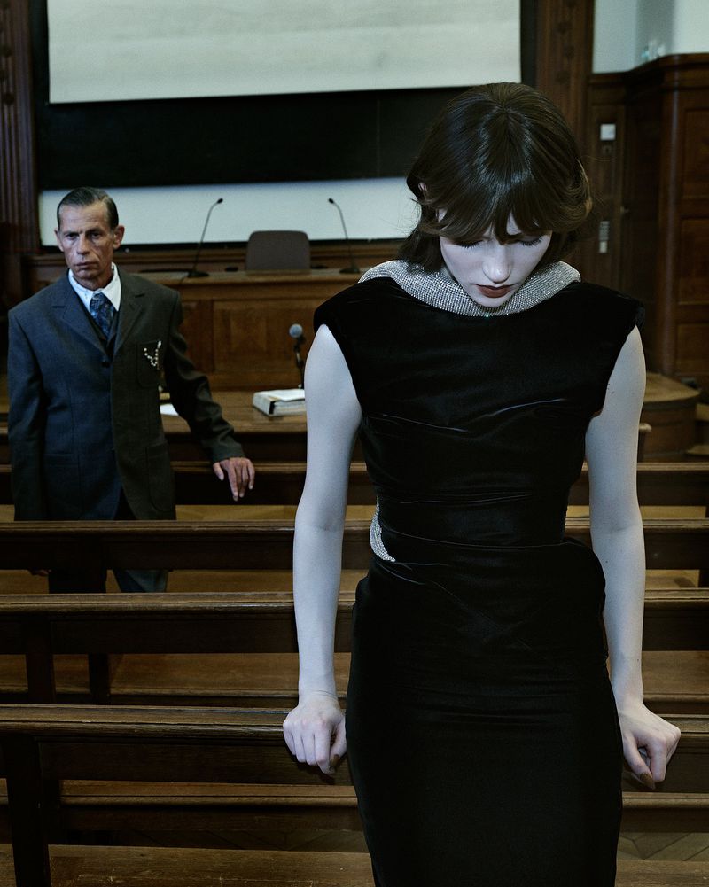Woman in a black dress standing in a courtroom next to wooden benches, with a man in a suit standing in the background.