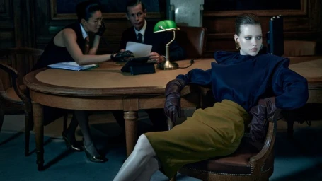 Olga Lane in a navy blouse, olive skirt, and leather gloves seated at a boardroom desk in a dimly lit office for Numéro France.