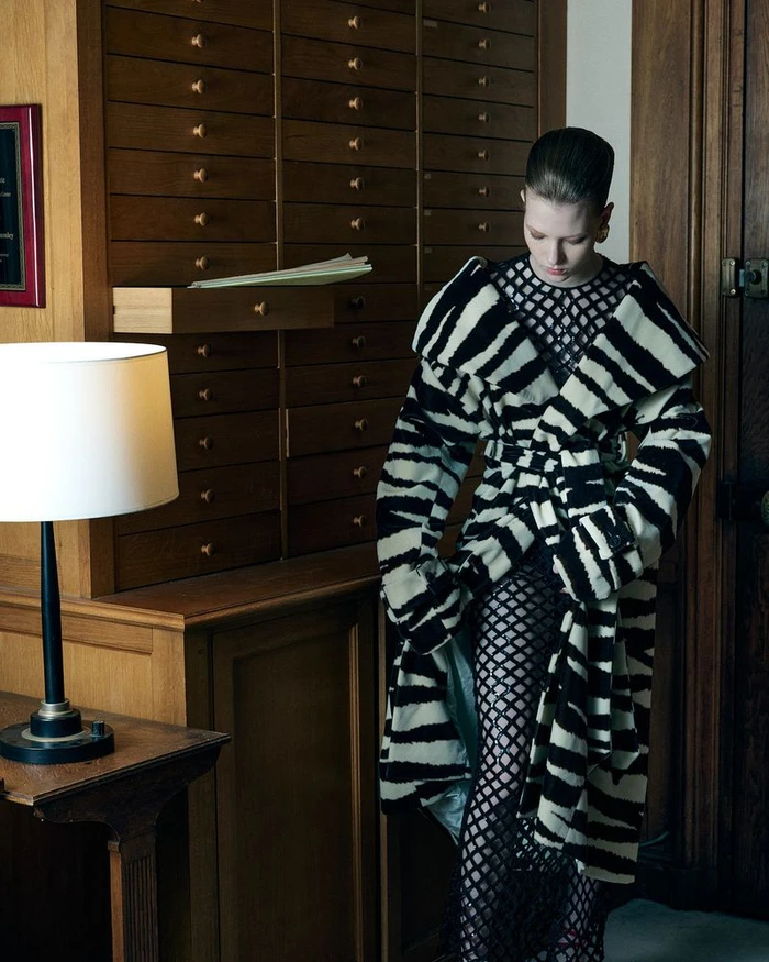 Model Olga Lane wearing a zebra-striped coat over a black fishnet jumpsuit standing beside a wooden filing cabinet.