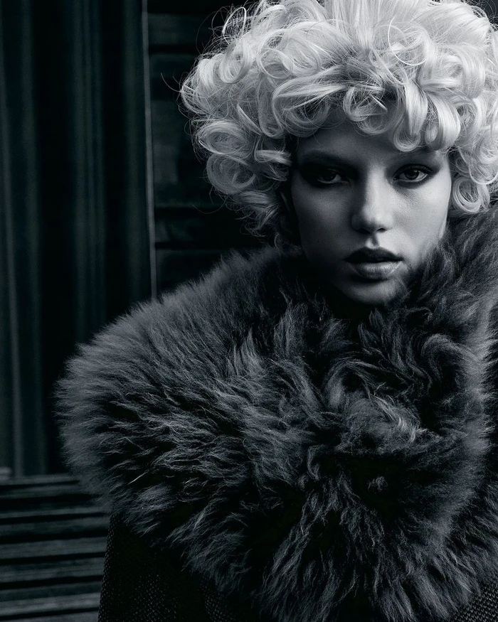 Monochrome close-up portrait of model Olga Lane with short curly blonde hair wearing a voluminous dark fur collar.
