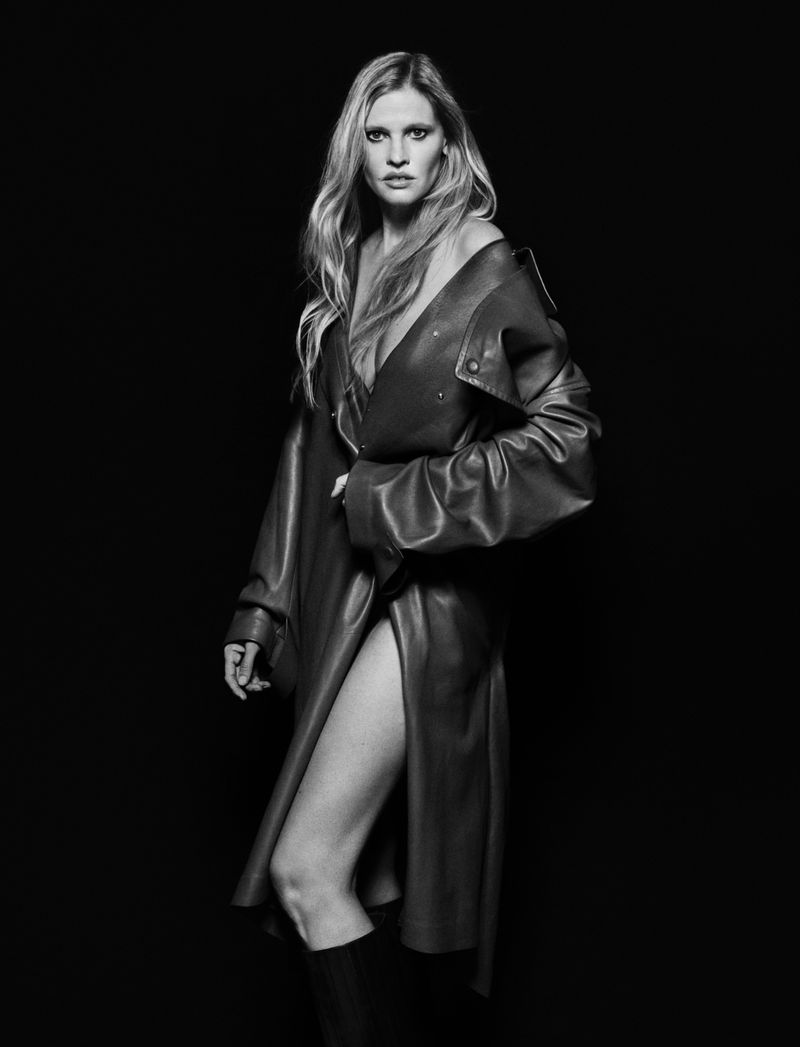 Black and white fashion shot of a woman with long blond hair wearing a leather coat and tall boots against a dark background.