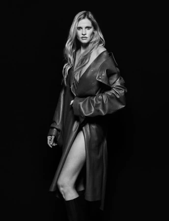 Black and white fashion shot of a woman with long blond hair wearing a leather coat and tall boots against a dark background.