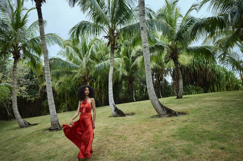 A woman in a red dress stands amidst a lush grove of palm trees, showcasing fashion in a tropical setting with a blurred background.