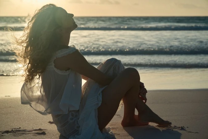 Adult female with curly hair sits on a sandy beach with a white dress enjoying the sunset over the ocean in a scenic wide shot.