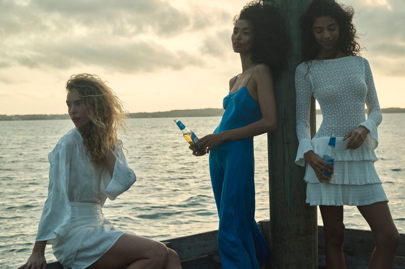 Three women in dresses pose by the sea at dusk, one holding a beer bottle. Soft light and open water create a serene backdrop.