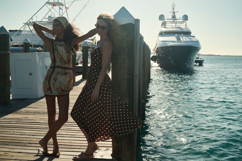 Two women in summer dresses pose on a wooden dock with yachts in the background on a sunny day, capturing a stylish outdoor scene.