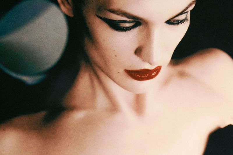 High-angle beauty portrait of a woman with bold winged black eyeliner and glossy red lipstick looking downward.