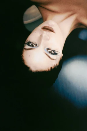 Close-up portrait of a woman with distinctive makeup looking at the camera, featuring dark hair and a dark background with blurred lights.