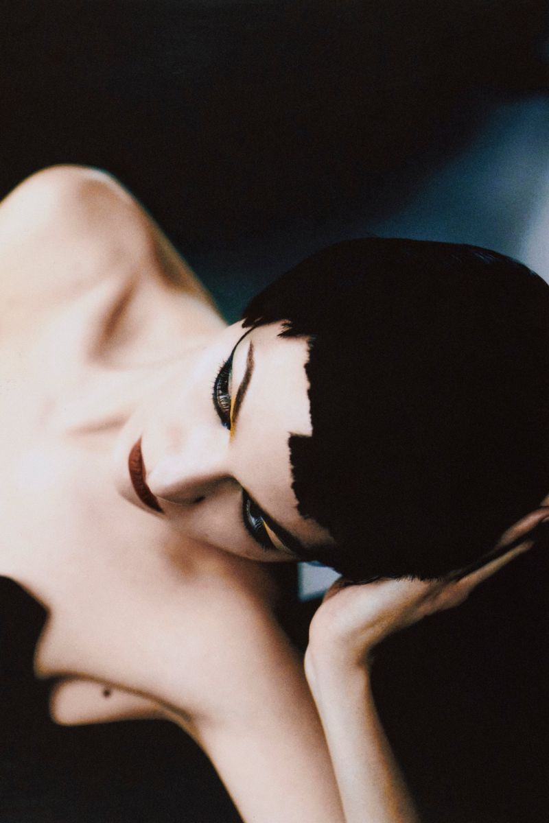 Artistic close-up of a woman with a dark bowl cut, dramatic eye makeup, and red lipstick, lying down in front of a dark background.