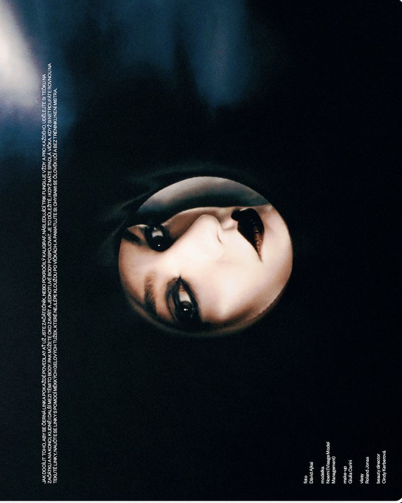 Fashion magazine cover with portrait of a pale-skinned adult female with heavy makeup, dark lipstick, and a shadowy black background.