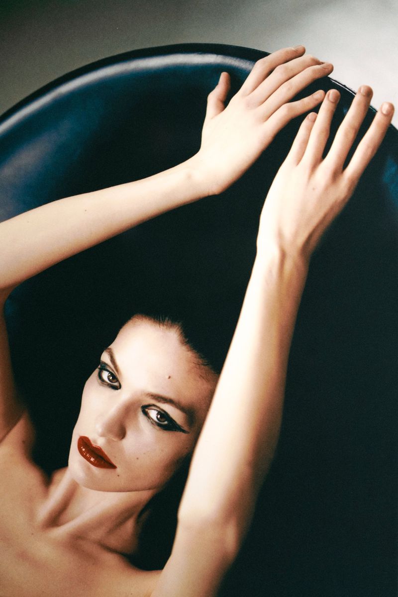 Portrait of a brunette woman with red lips lying in a black chair, hands raised above her head in a high-key photo studio shot.