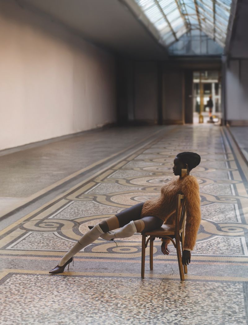 Fashionable woman with stylish haircut poses on a chair in a long corridor with a patterned floor and glass ceiling for editorial content.