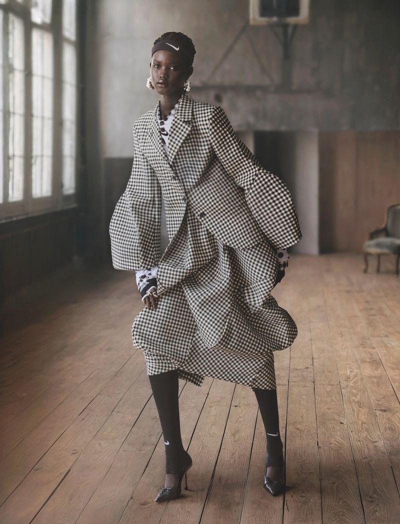 Elegant adult female fashion model poses in a checkered blazer and skirt with black tights and heels in an indoor studio setting.