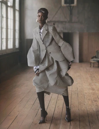 Elegant adult female fashion model poses in a checkered blazer and skirt with black tights and heels in an indoor studio setting.