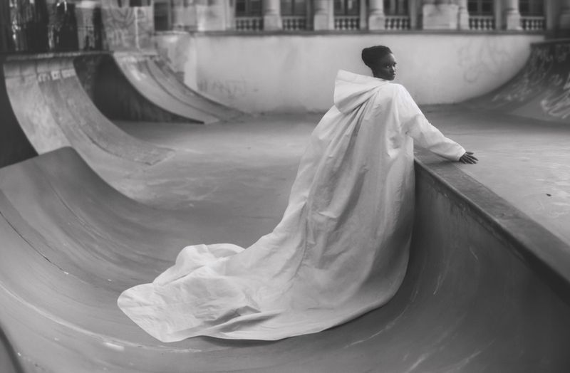 Black-skinned model poses in a flowing white hooded cape at an empty skate park, creating a striking contrast and fashion-forward image.