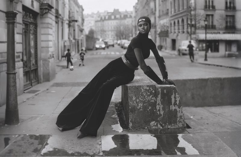 An adult female fashion model sits on concrete city street bollard in black sportswear with children playing soccer in the background.